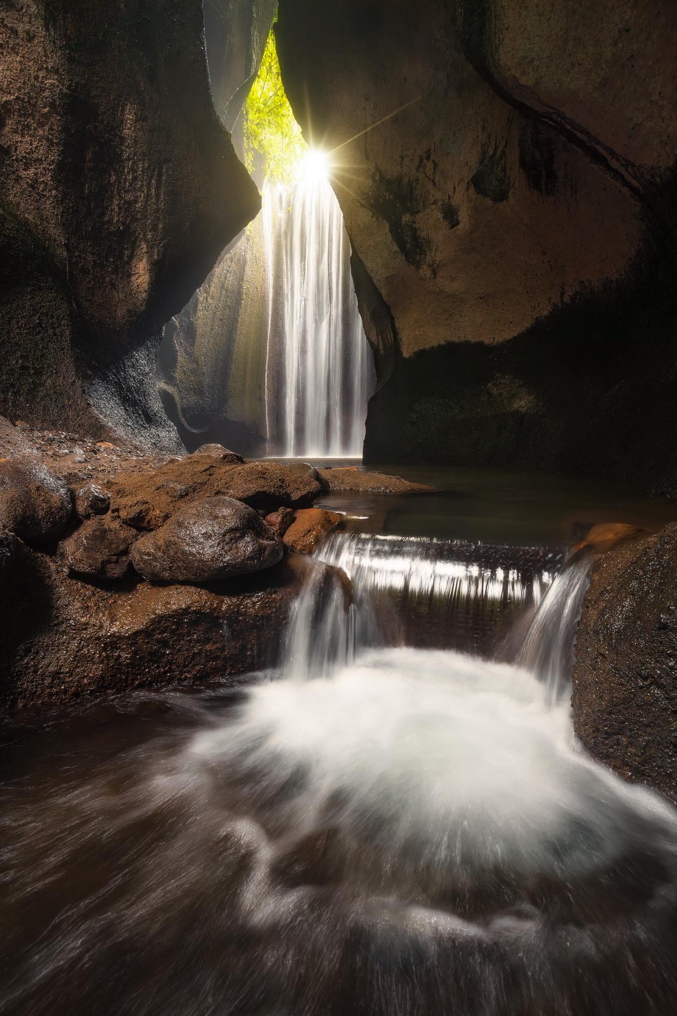 Sunlight shining into a small cave with a waterfall flowing into a pool surrounded by rocks.