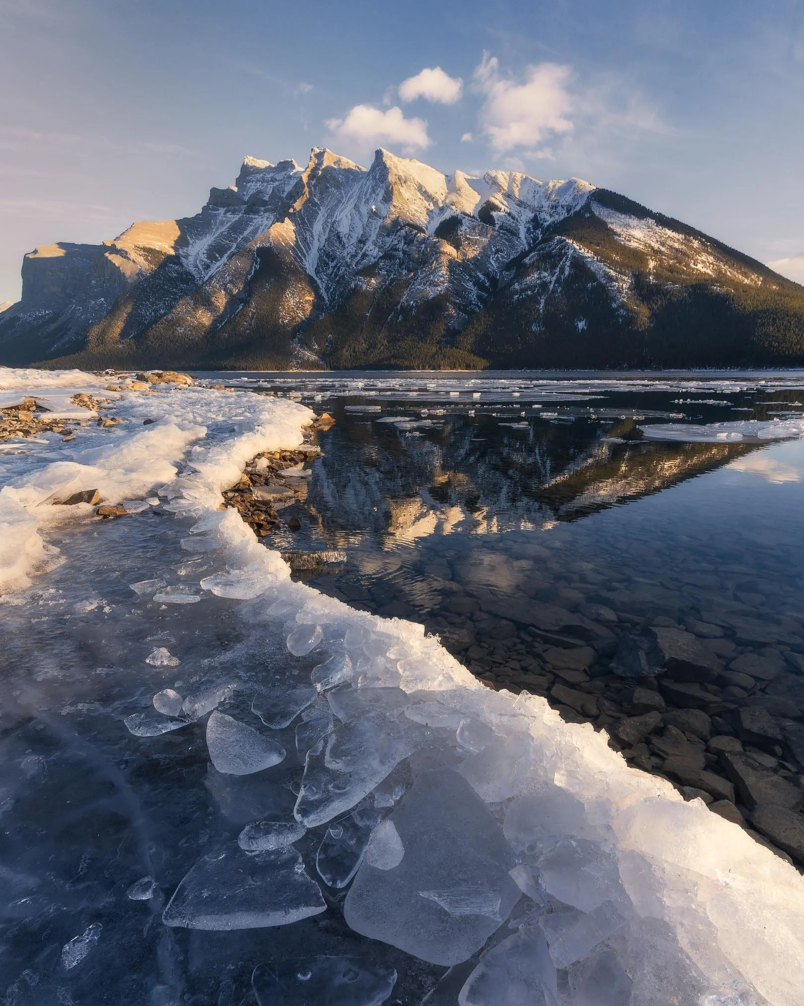 Snow-covered mountains, partially frozen lake, rocky shoreline, with clouds and blue sky overhead.