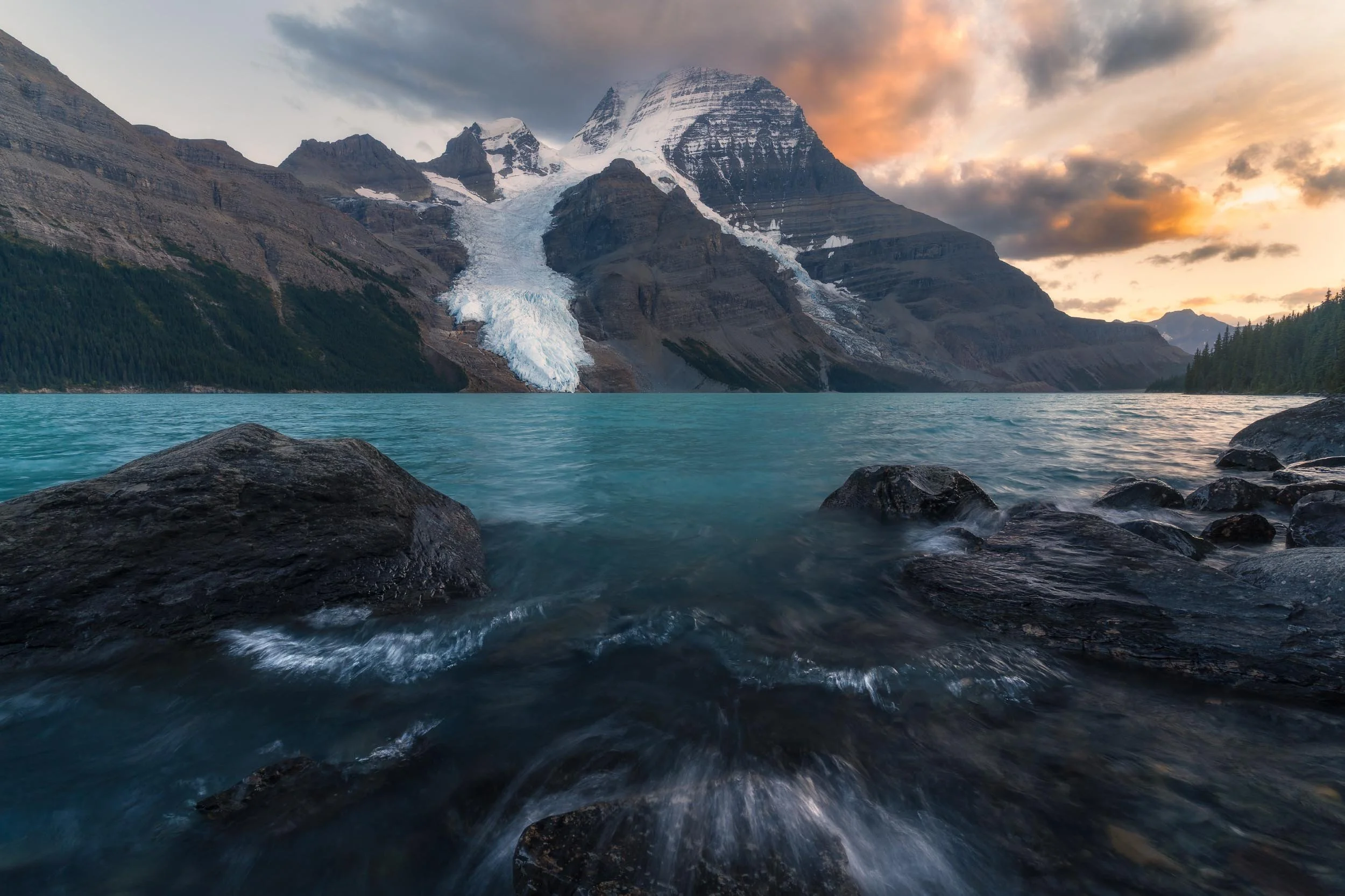A mountain with snow-capped peaks and a glacier flowing down its side, reflected in a turquoise lake at sunset, with dark rocks in the foreground and clouds in the sky.