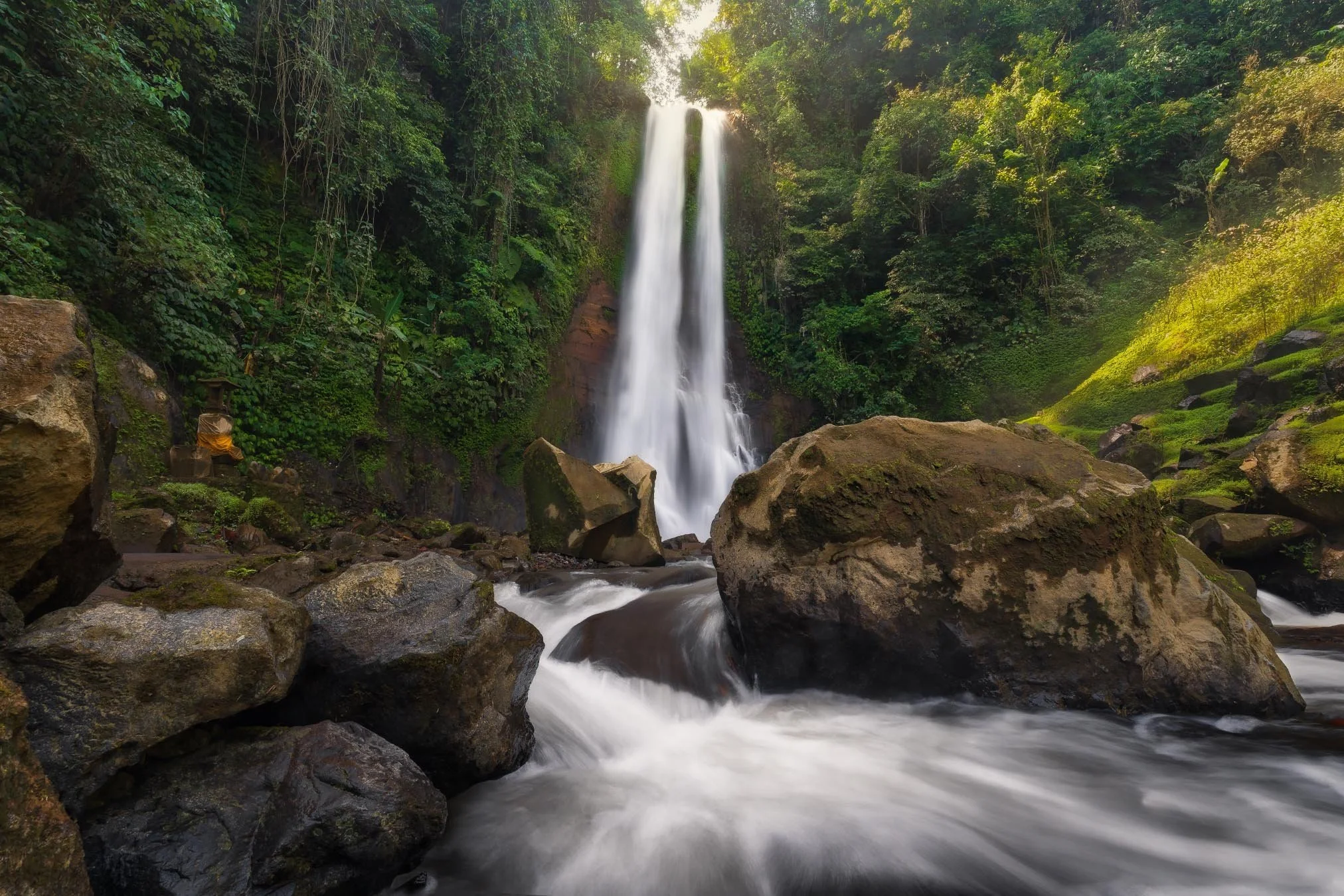 A tall waterfall cascading down into a rocky stream surrounded by lush green forest.
