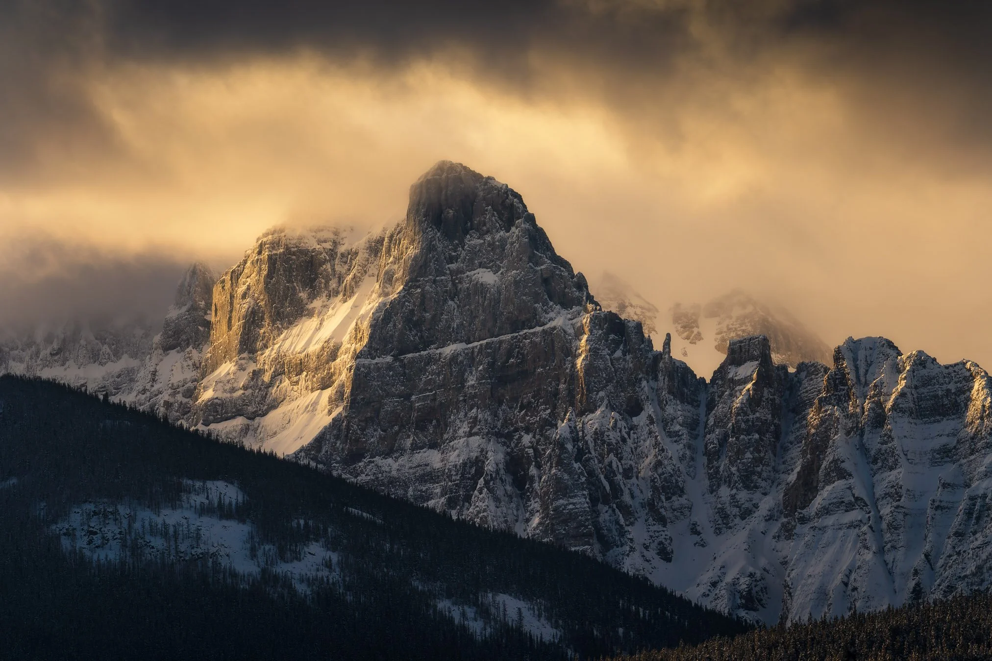 Snow-covered mountain peaks shrouded in clouds with a forested base in the foreground.