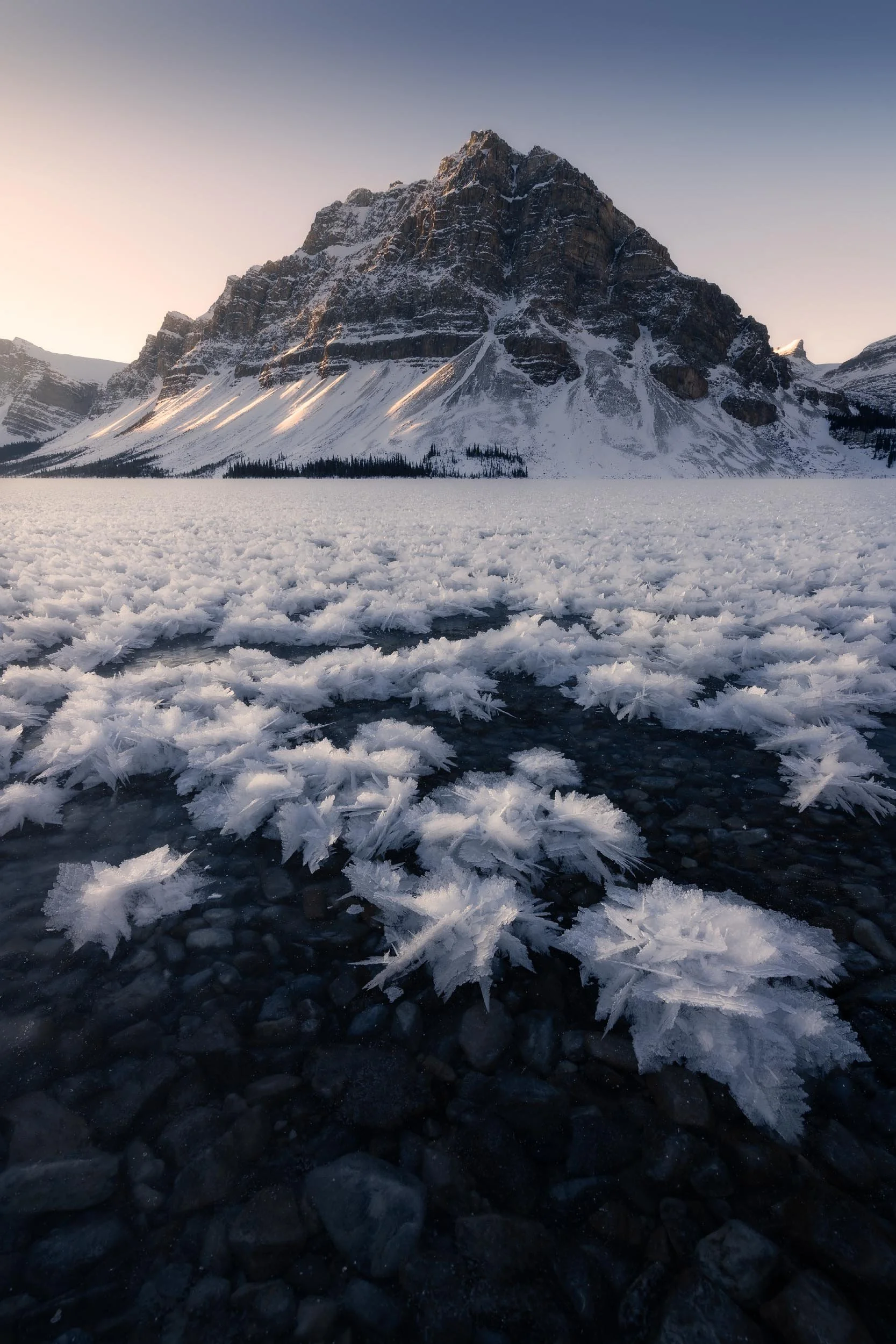 Snow-covered lake with ice formations in the foreground, towering snow-dusted mountains in the background, and a clear evening sky.