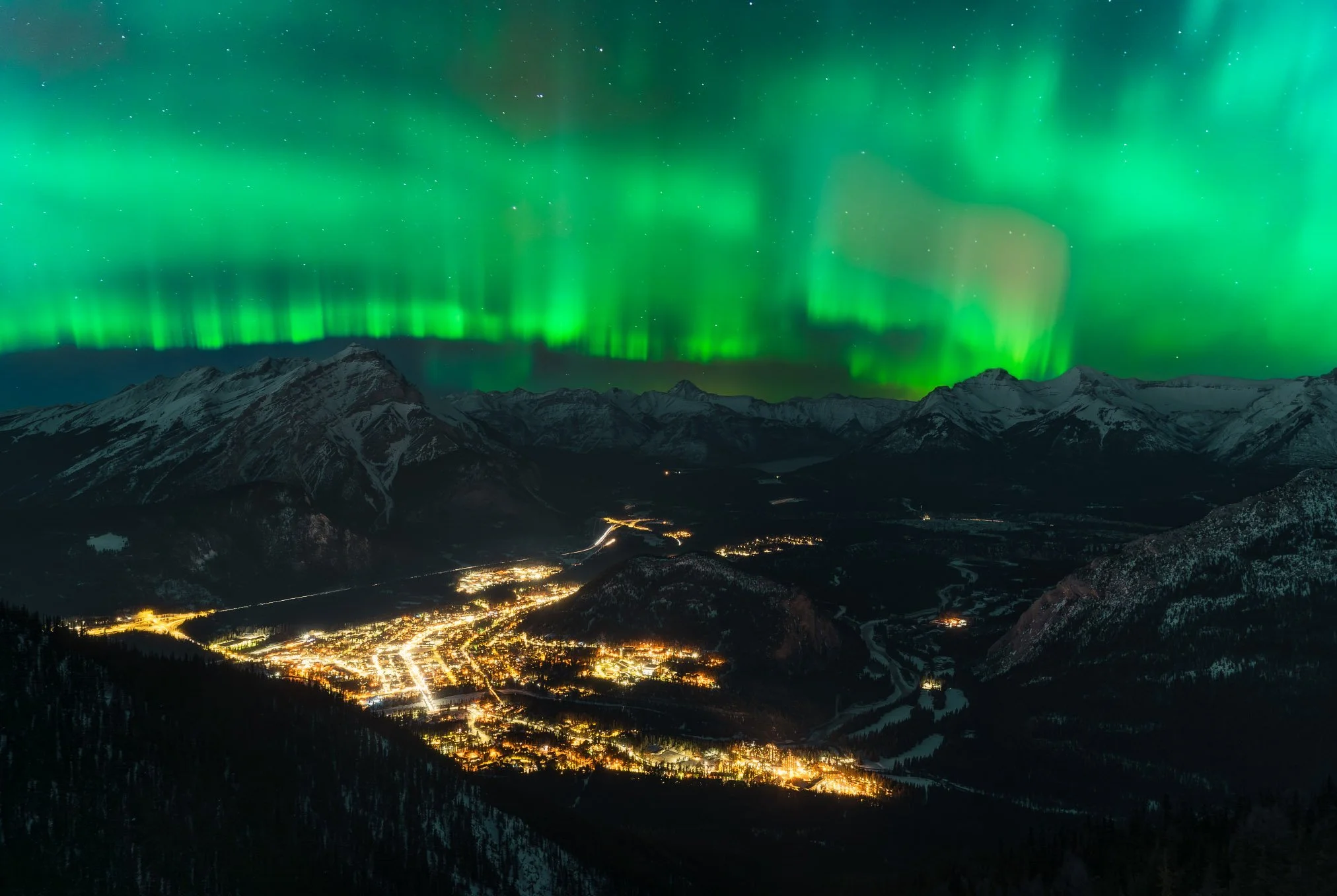 Northern lights glow green above mountains and a small town at night.