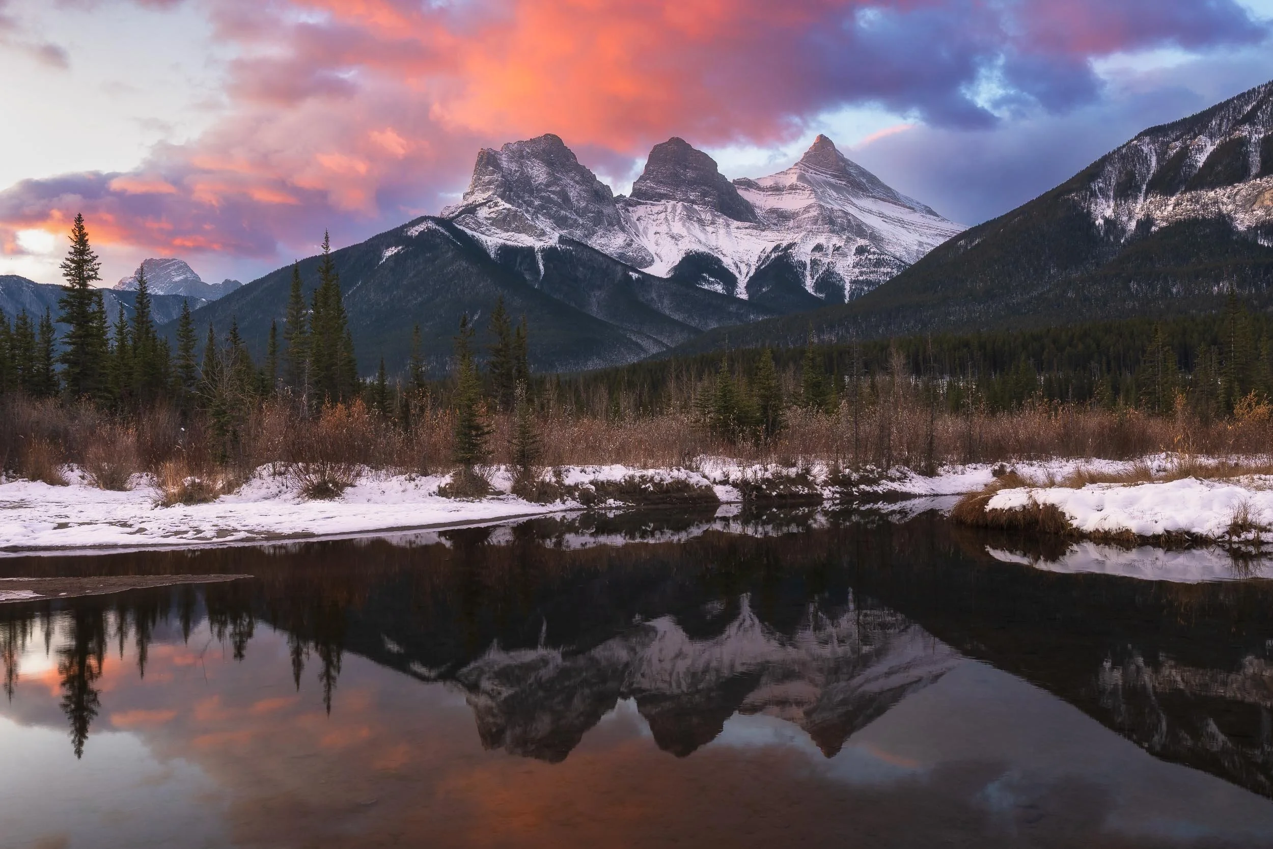 Snow-capped mountains against a colorful sunset sky reflected in a calm river, with a forest of evergreen trees in the foreground.
