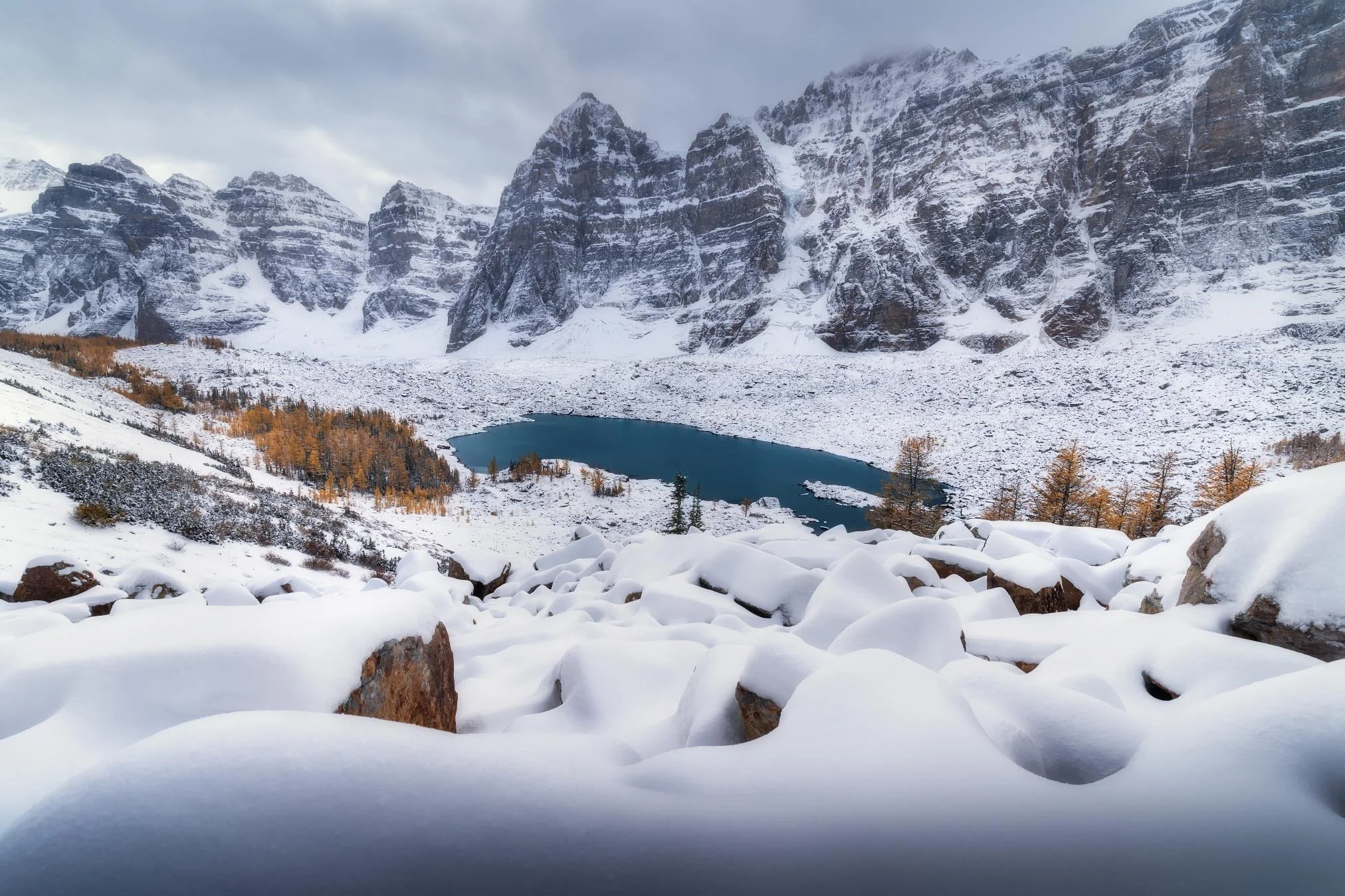 Snow-covered mountains with a small lake at the base, partially frozen, and snowy terrain with sparse trees in the foreground.