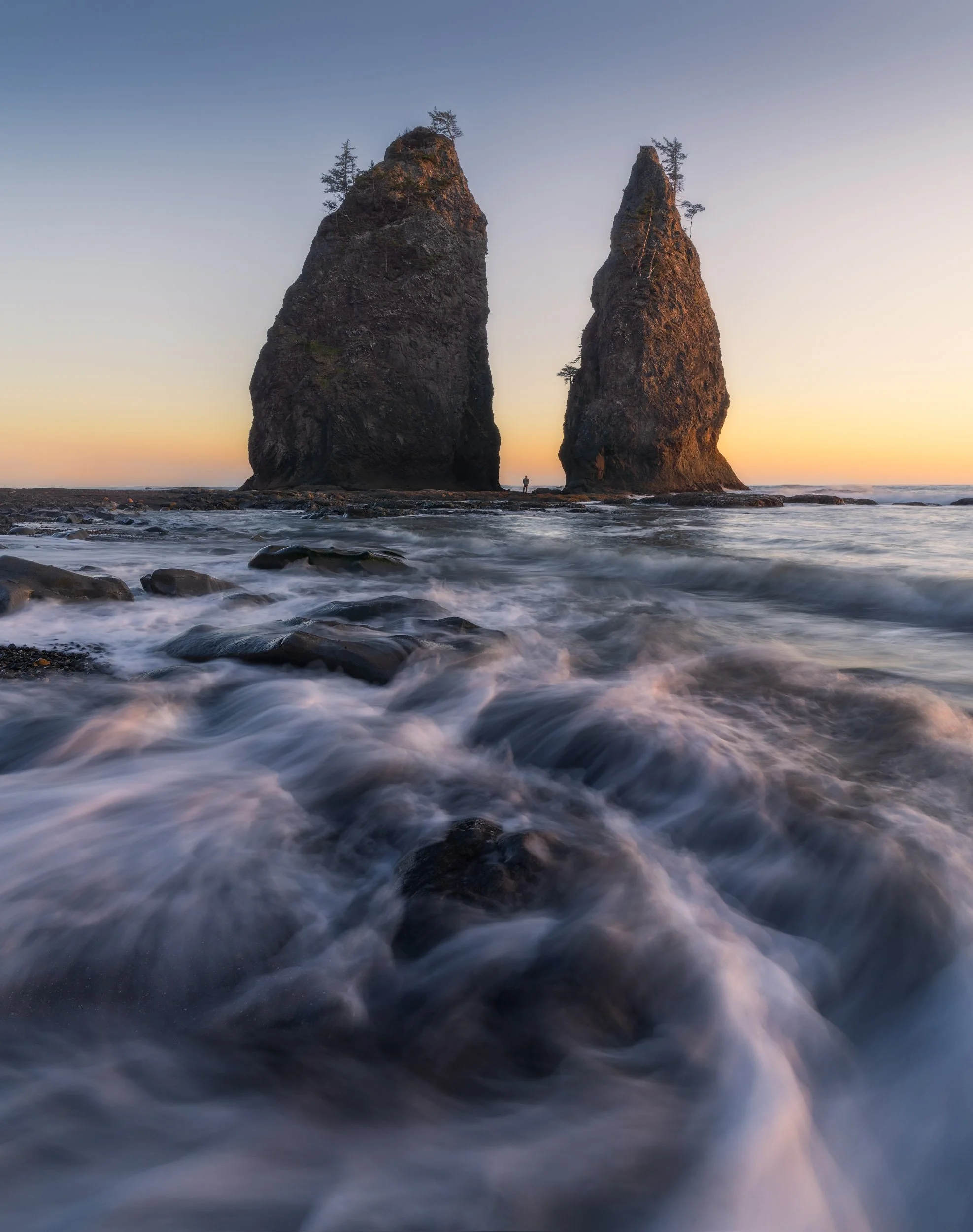 Two large rock formations in the ocean at sunset with trees growing on top, and waves crashing at the base.