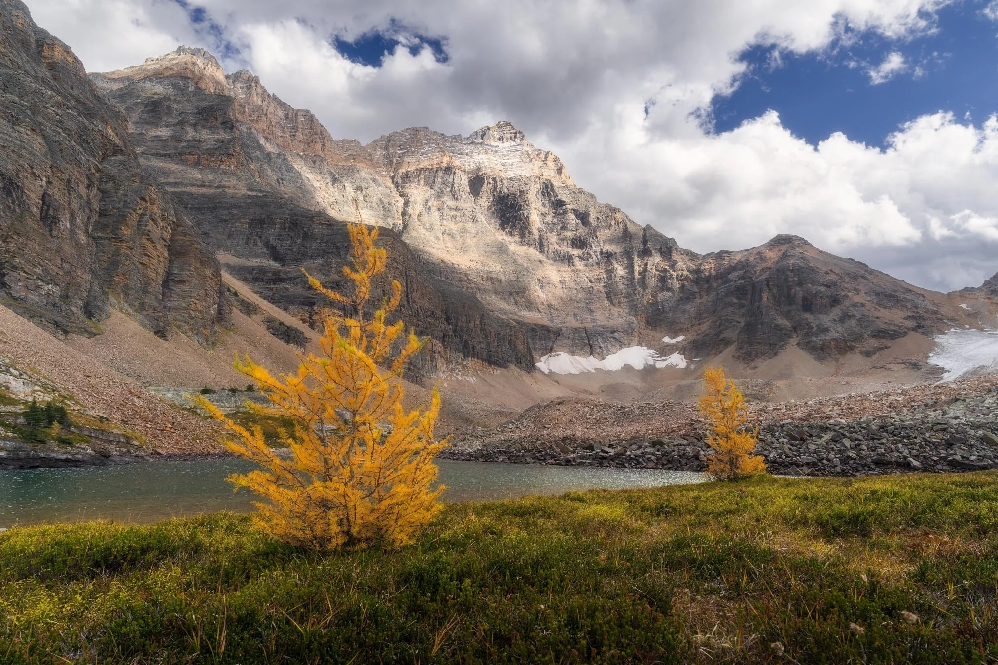 A mountain landscape featuring rocky, rugged mountains with patches of snow, a lake in the foreground, and yellow trees with cloudy sky overhead.
