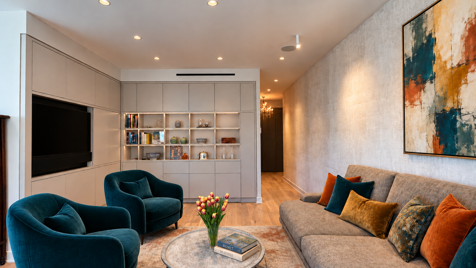 Living room with beige walls, a gray sofa with colorful pillows, two blue velvet armchairs, a round coffee table with a bouquet of tulips, and wall art. Built-in shelving with books and decor, TV, and hardwood floors.