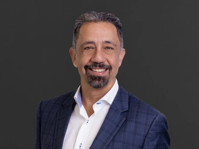 A smiling man with short gray hair and a beard, wearing a blue checkered blazer and white shirt, standing against a dark gray background.