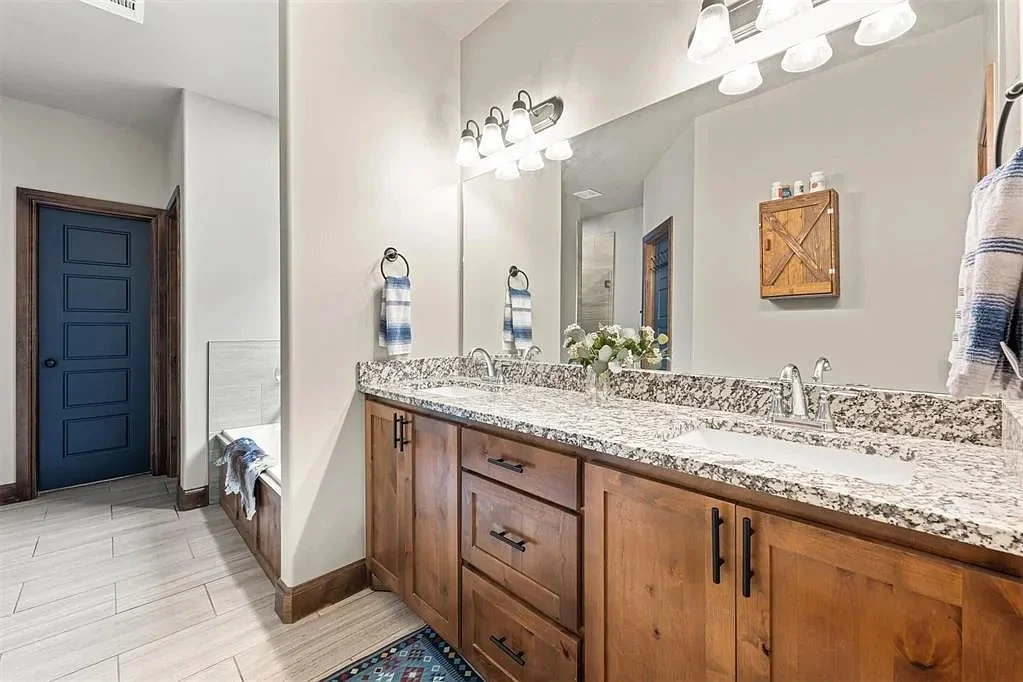 Bathroom with a double sink granite countertop, wooden cabinets, mirror, wall-mounted light fixtures, towel hooks with towels, a small wooden cabinet on the wall, and a door painted blue.