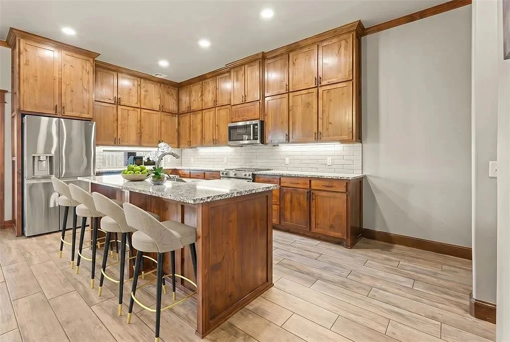 Modern kitchen with wooden cabinets, stainless steel refrigerator, microwave, and a kitchen island with bar stools, next to a light-colored tiled backsplash.