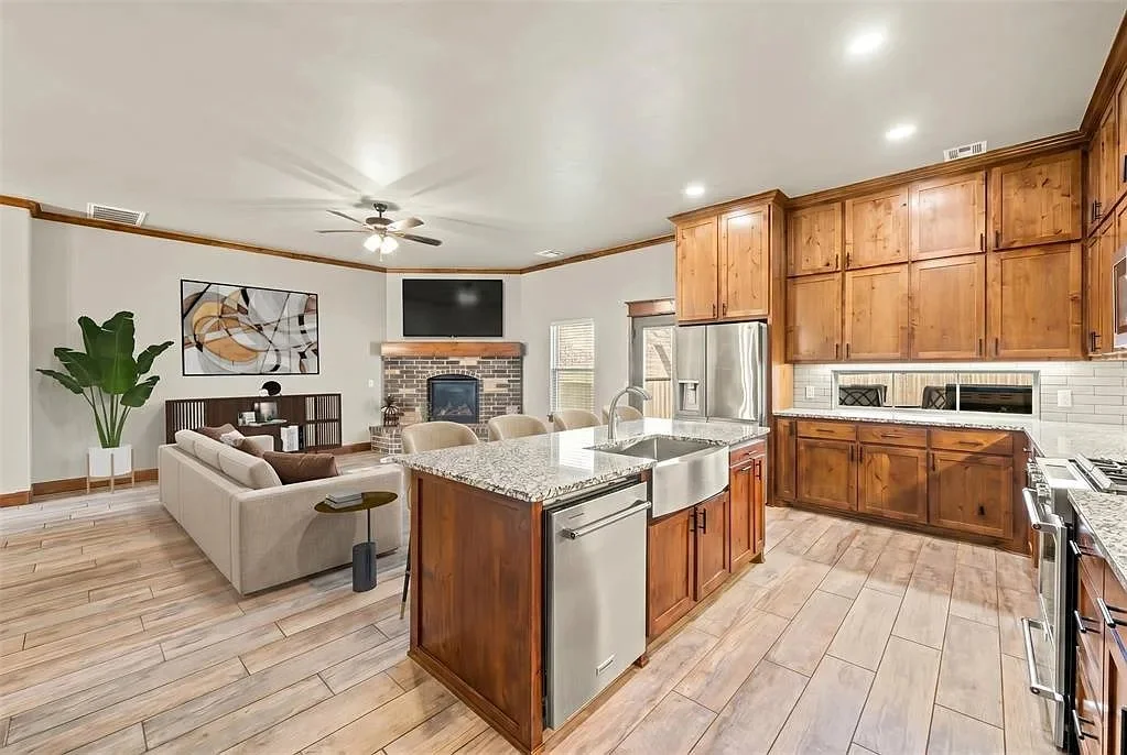 Open-concept kitchen and living room with wooden cabinets, granite countertops, and a brick fireplace, featuring a ceiling fan and large windows.