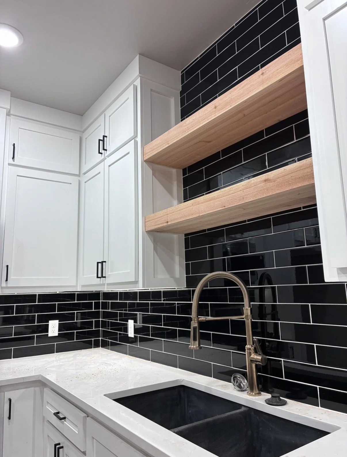 Modern kitchen with white cabinets, black tile backsplash, and two wooden open shelves. Silver faucet over a black sink and white countertops.
