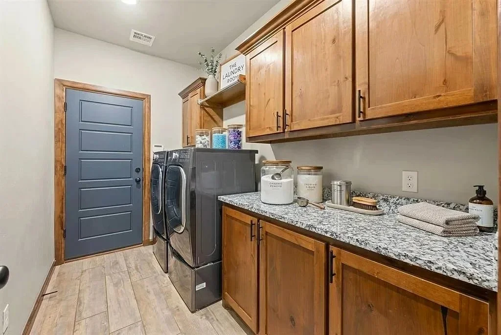 A laundry room with a blue door, wooden cabinets, a granite countertop, and a black washing machine and dryer. The counter has containers, a towel, and a soap dispenser. There is a decorative sign and plants on top of the cabinets.