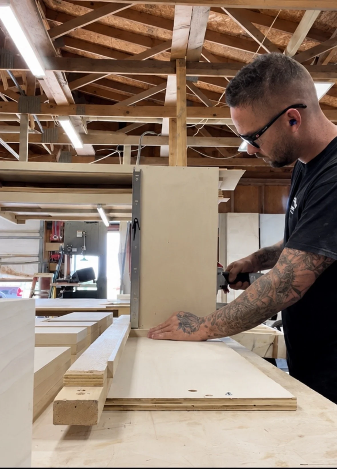 A man with tattoos on his arms wearing glasses with ear protection is working in a custom cabinet shop. He is using a power tool on a piece of wood, with additional cabinet pieces and workshop tools visible around him.