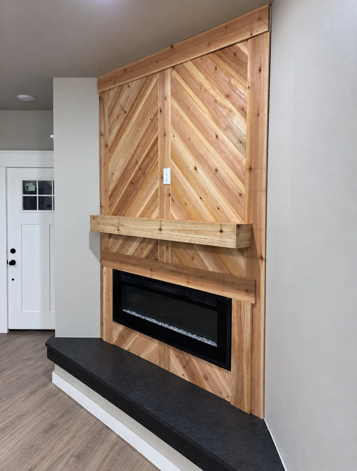 A modern living room fireplace with a chevron-patterned wooden surround and a black fireplace insert, accented by a wooden mantel, positioned against a light colored wall.