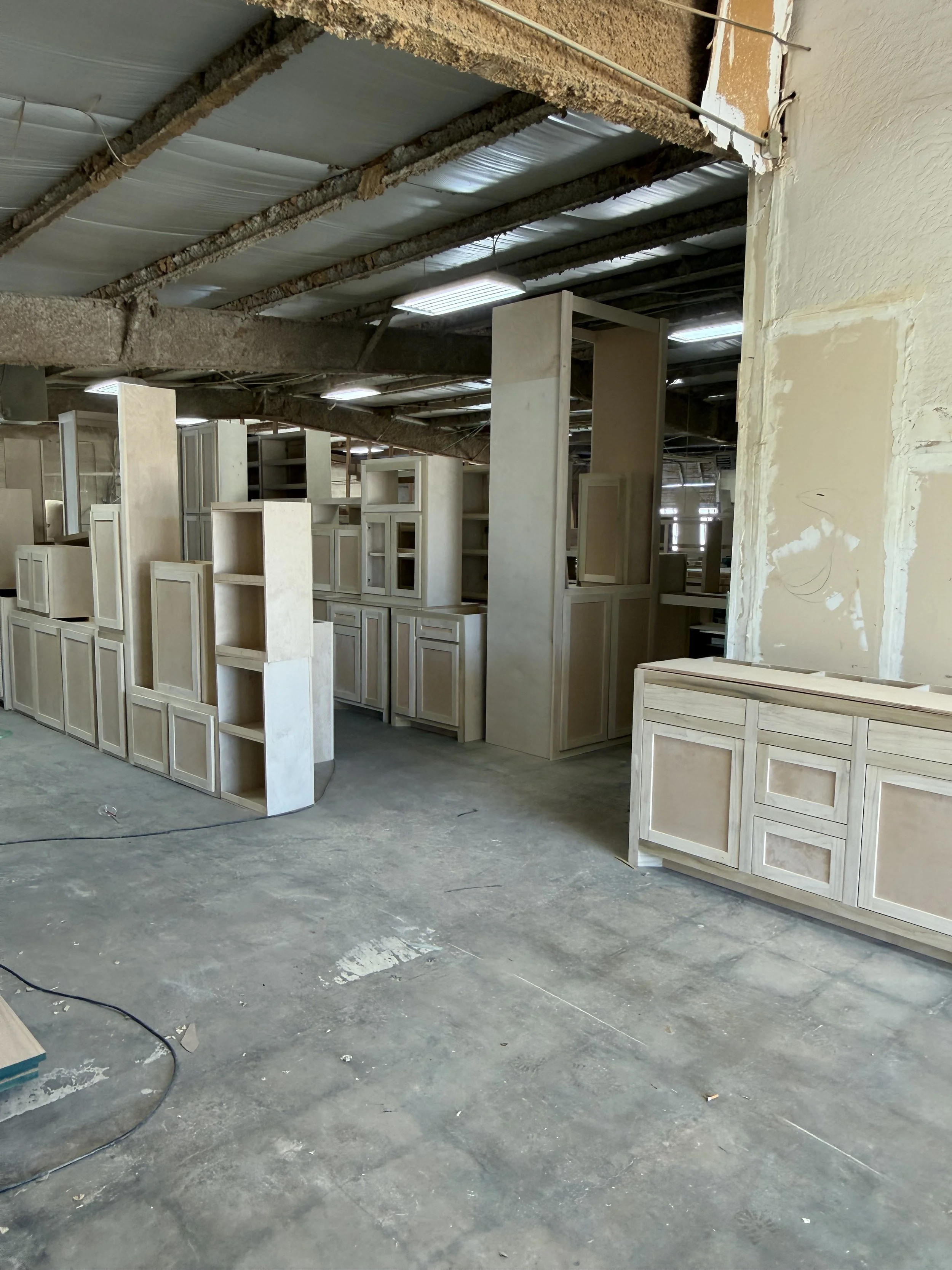 Inside custom cabinet workshop, showing wooden cabinetry and shelving units, concrete floor, and construction materials.