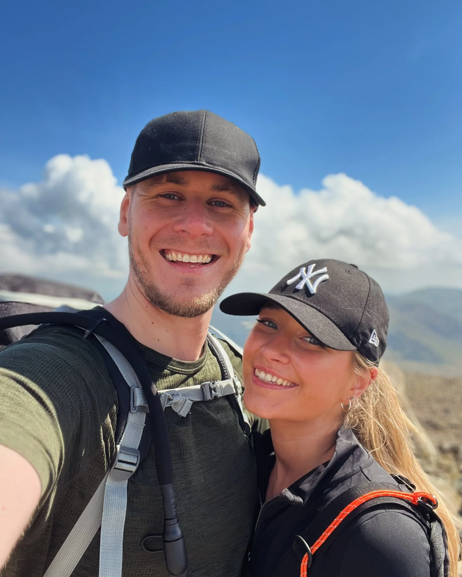 Close-up of smiling man and woman wearing black caps and outdoor clothing, standing together outdoors with a blue sky and clouds in the background.