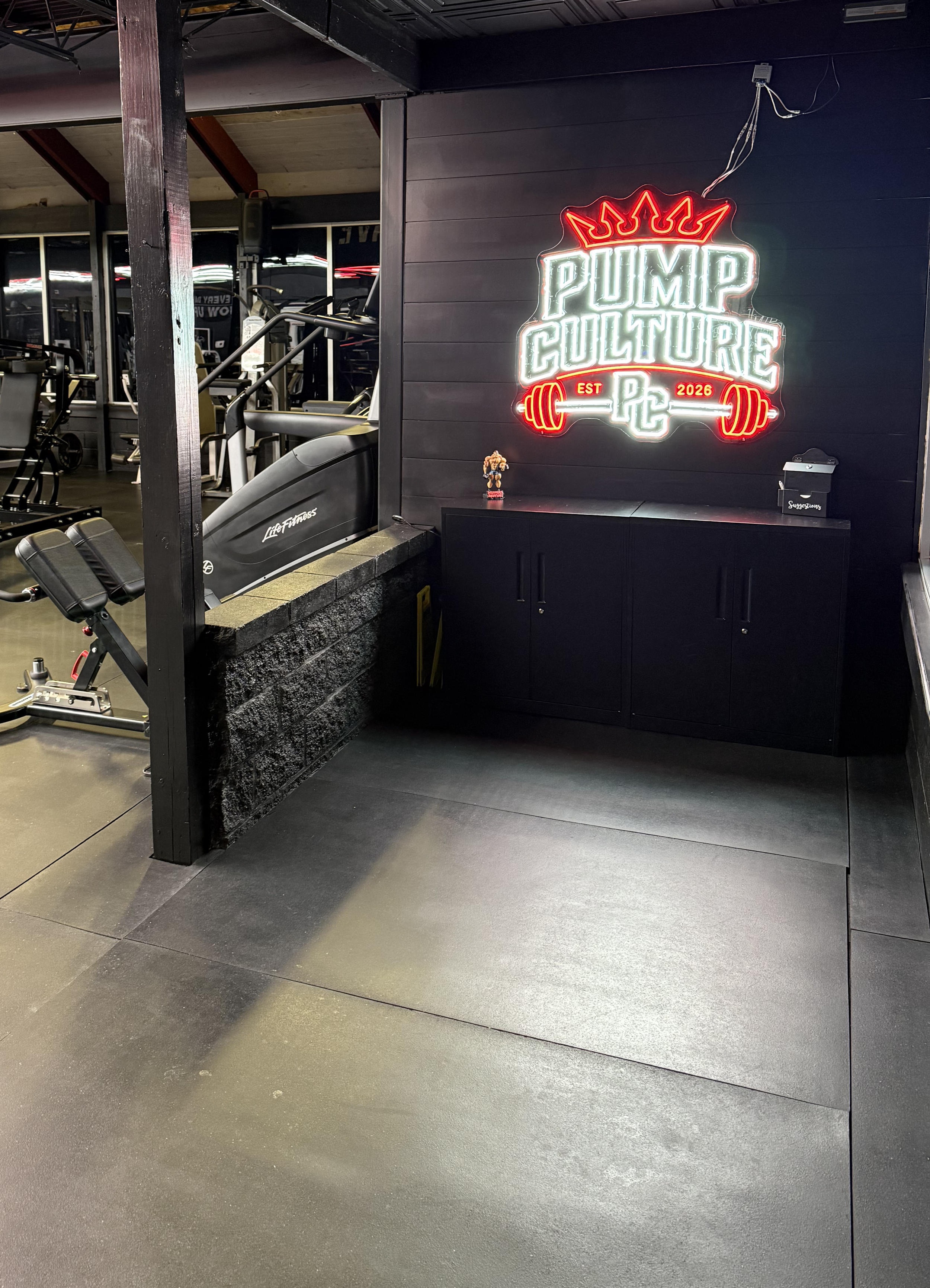 A fitness gym with exercise equipment on the left side, including ellipticals and stationary bikes. On the right side, there's a black cabinet with a small muscular figurine, a box labeled 'Suggestions,' and a large illuminated sign that reads 'Pump Culture' with a crown above and barbell with weights below, indicating it was established in 2026.