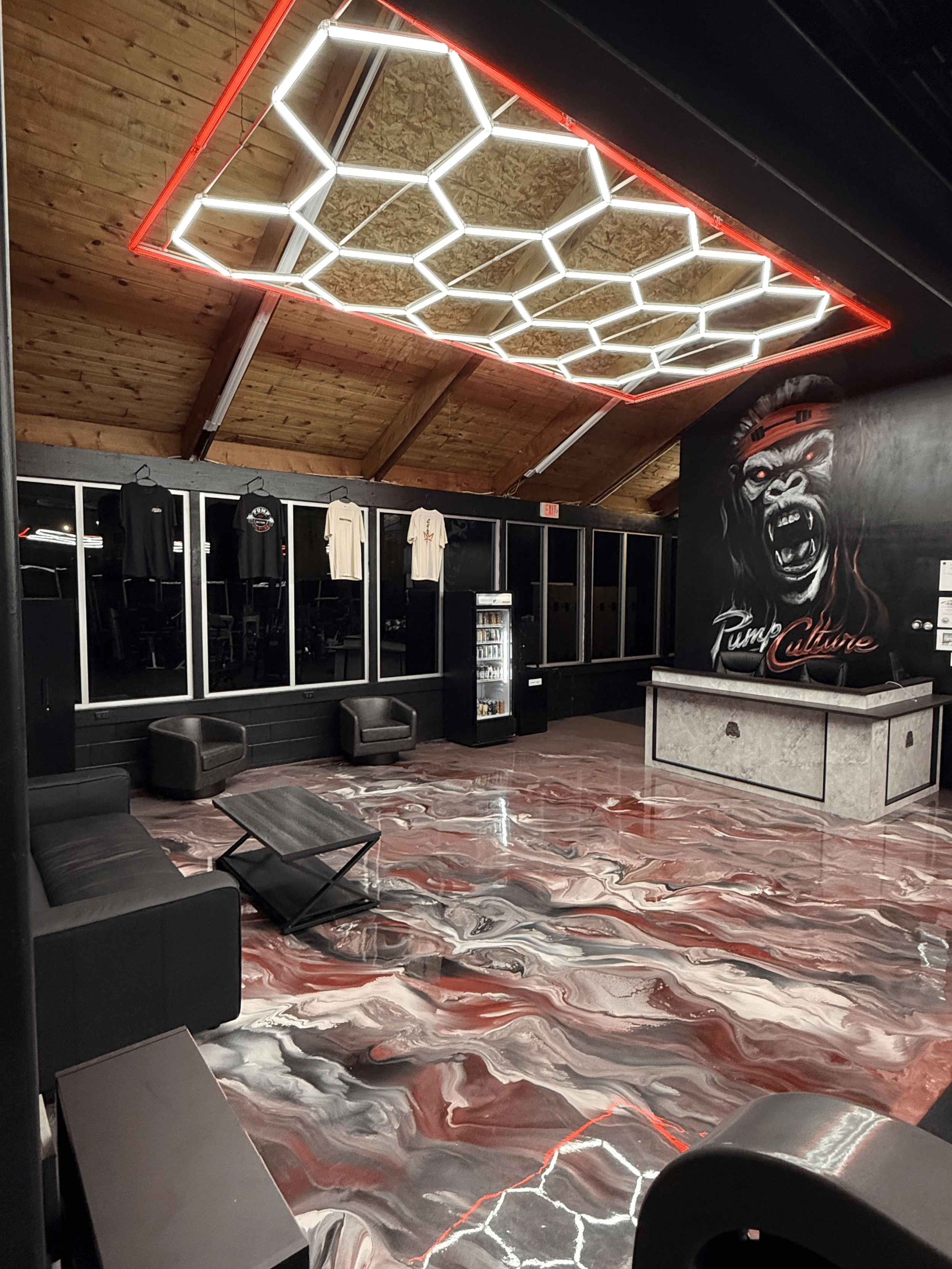 Interior of a gym with a wooden ceiling, black walls, and a colorful marble floor. LED hexagon ceiling lights and a large mural of a roaring gorilla with the text "Pump Culture" are visible.