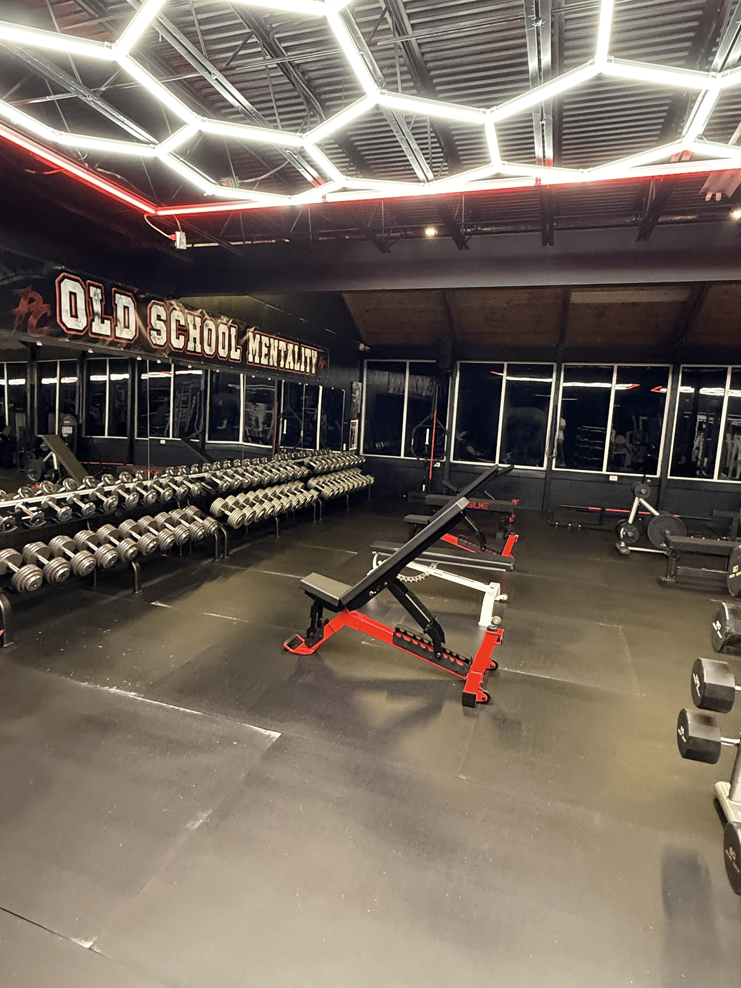 Empty gym with dumbbells on racks, workout benches, and fitness equipment, with decorated sign reading 'Old School Mentality' on the wall. Overhead hexagonal lighting fixtures illuminate the space.