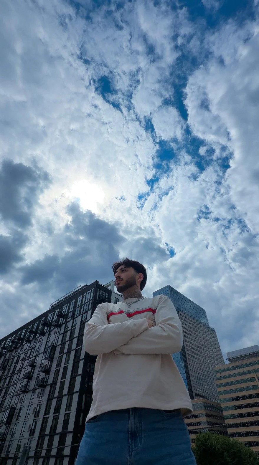 A young man standing outdoors with arms crossed, looking to the side, in front of tall buildings under a partly cloudy sky.