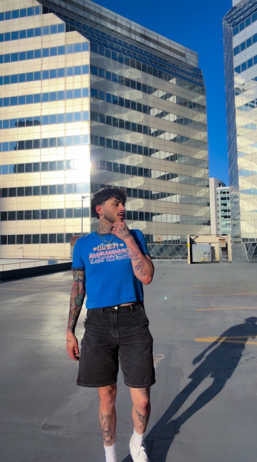 A young man with tattoos standing on a rooftop parking lot in front of modern glass buildings under a blue sky.