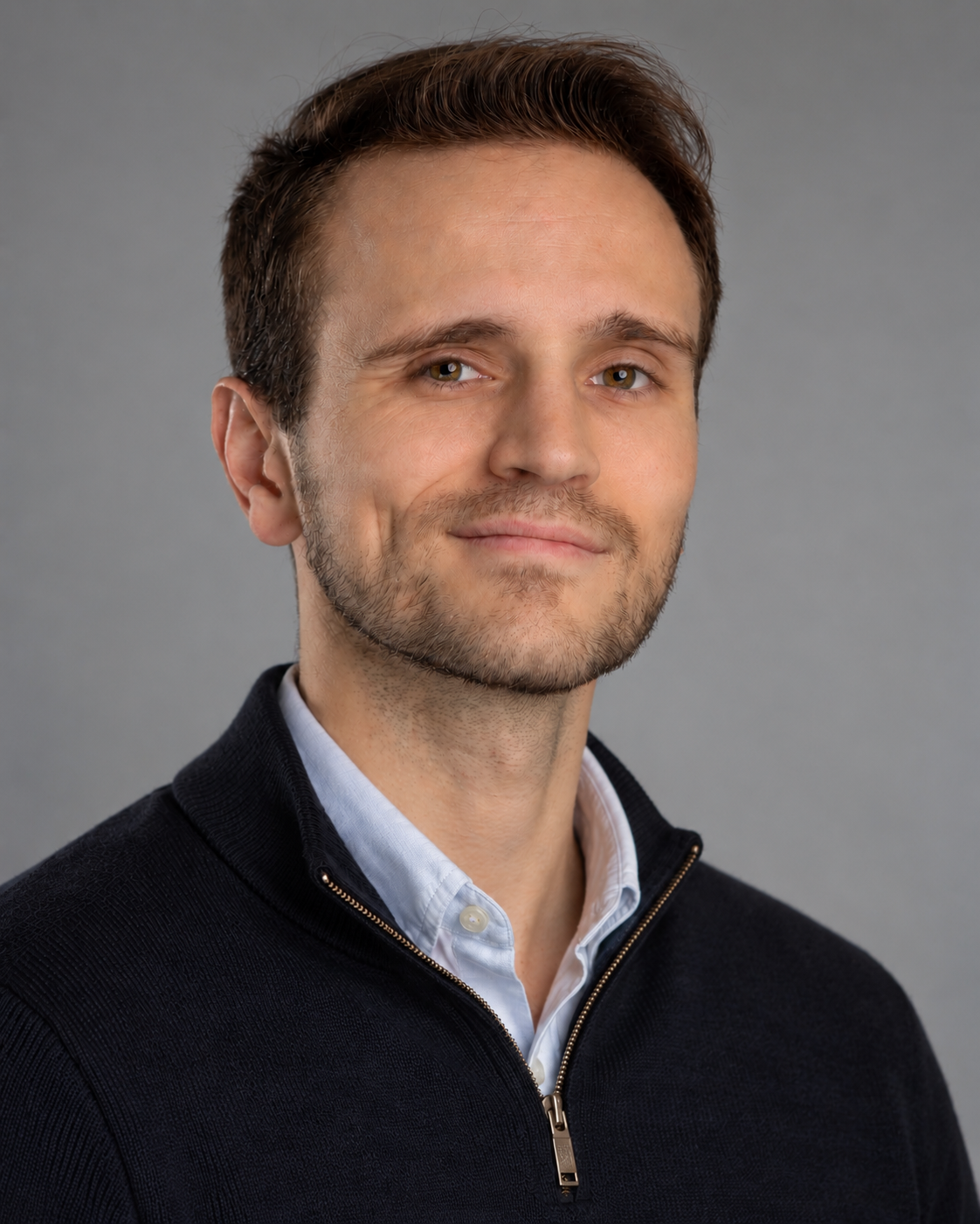 A man with light skin, short brown hair, and slight facial hair, wearing a black zip-up sweater over a collared shirt, smiling softly at the camera, against a plain gray background.