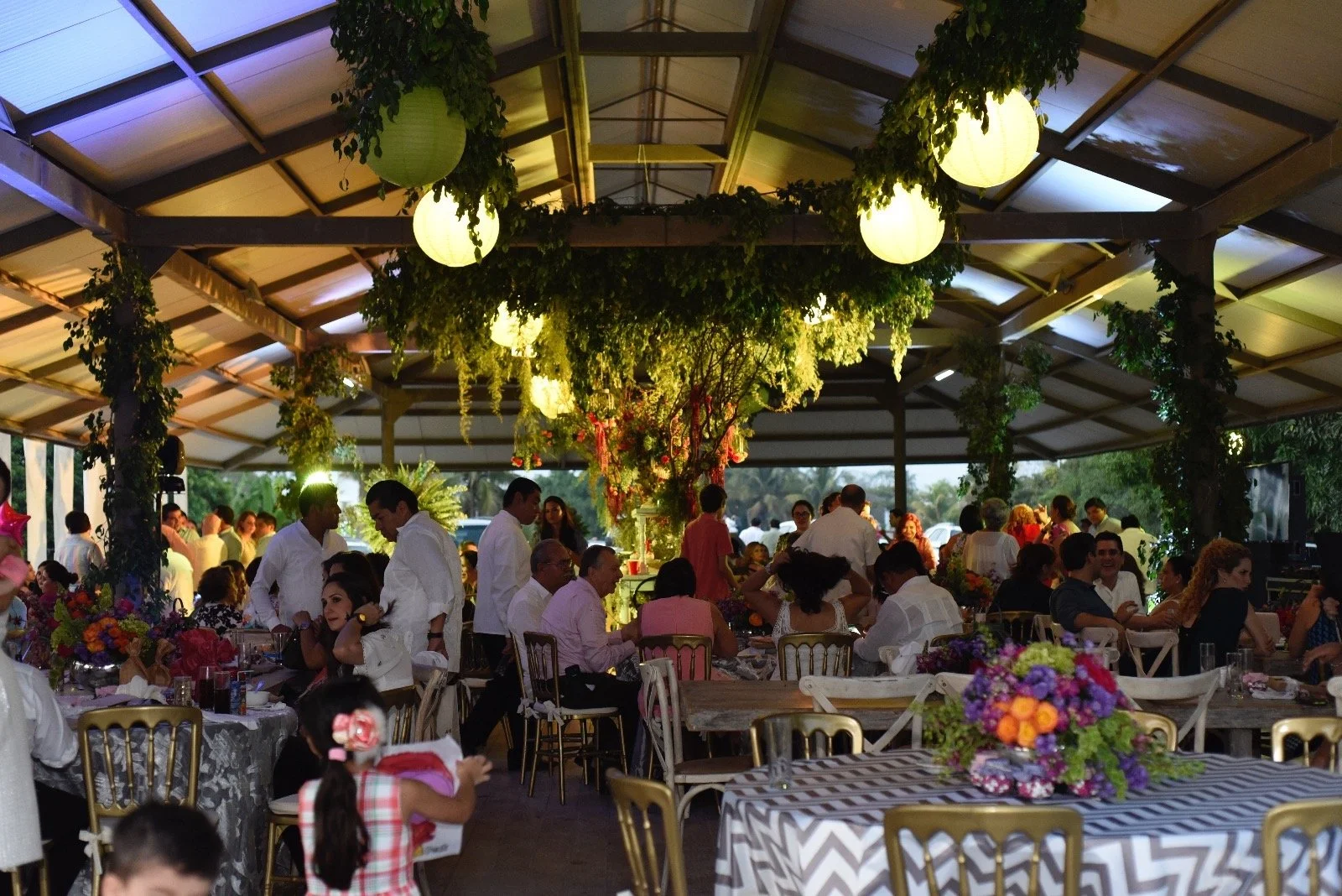 Una celebración o evento social al aire libre en un espacio techado con celebrantes y decoraciones florales y de papel de colores.