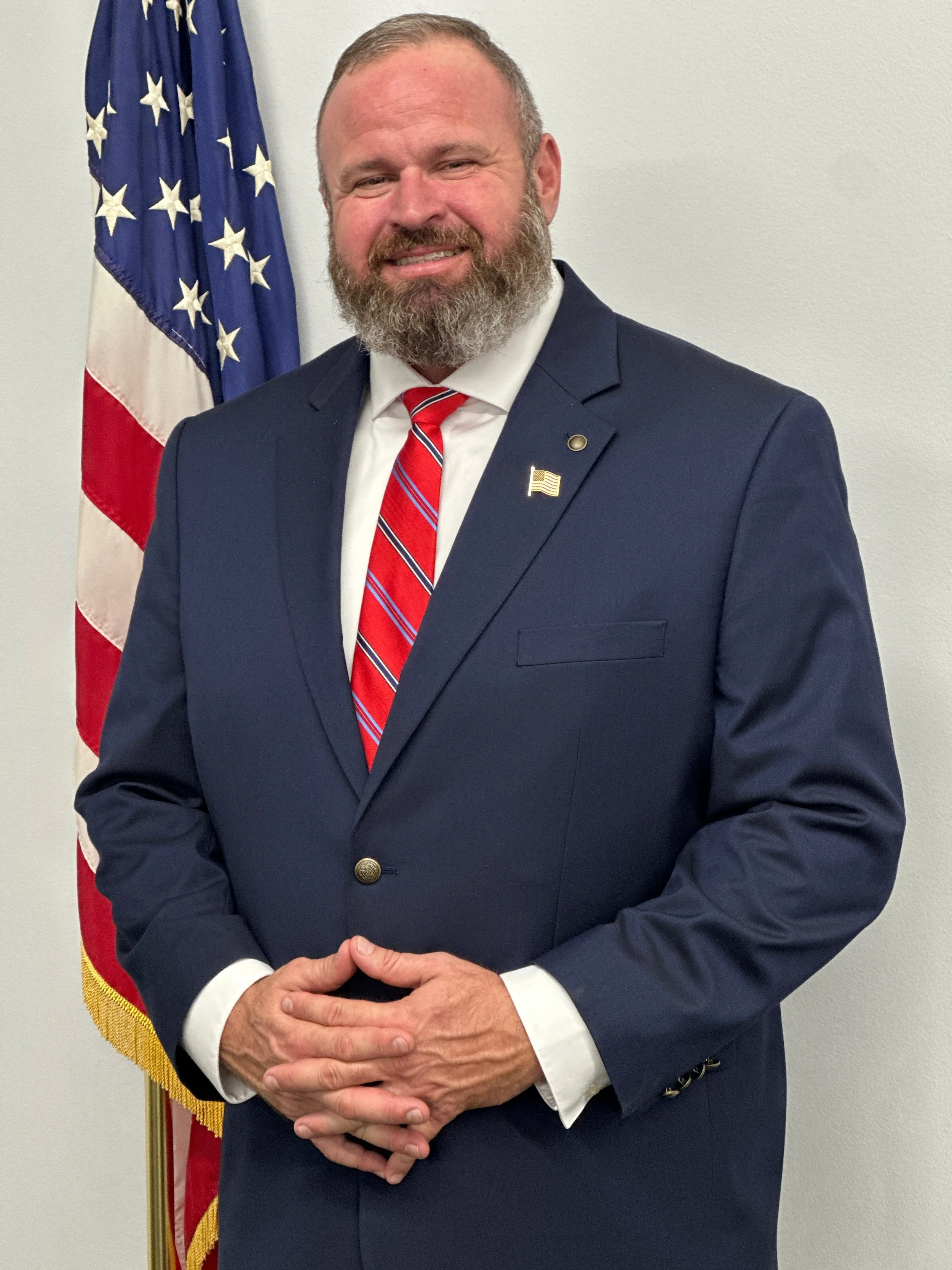 A man in a navy suit with a white shirt and a red tie with blue and white lines, standing in front of an American flag. He has a beard, short hair, and is smiling. He has a small American flag lapel pin on his suit jacket.
