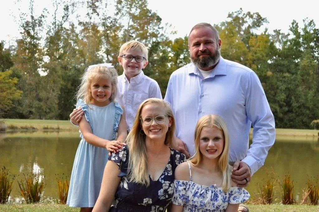 Family portrait of six people outdoors by a lake, including two adults and four children, with trees and water in the background.