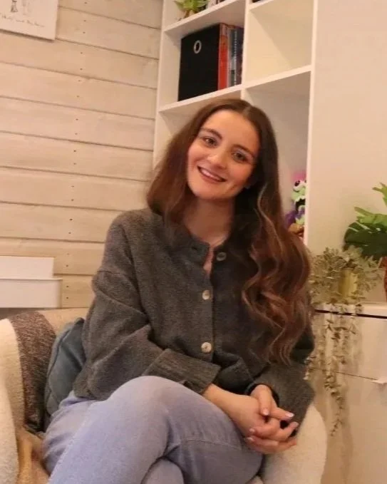 A young woman with long brown hair smiling while sitting on a beige couch in a cozy room with wooden walls and white shelves filled with books and decorative items.