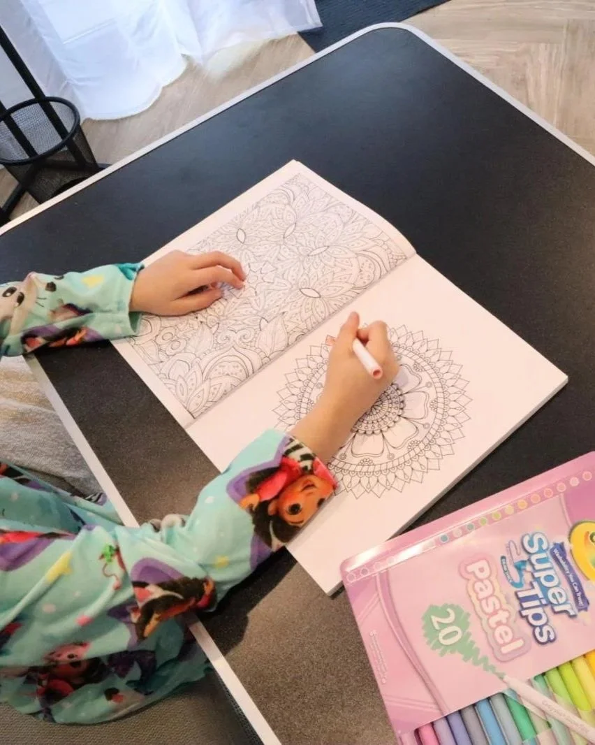 Child coloring intricate mandala patterns in a coloring book with markers on a black table.