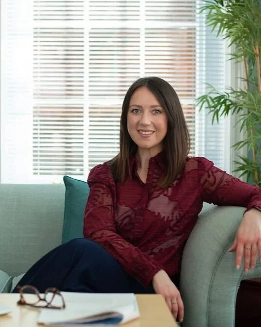 A woman with shoulder-length dark hair sitting on a light-colored couch in a bright room with window blinds and a large green plant in the background.