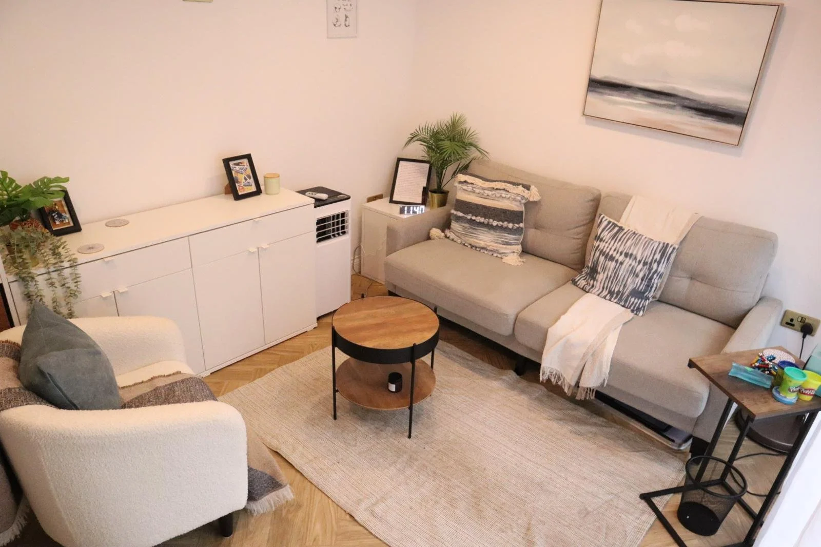 A cozy living room with a white armchair, a beige sectional sofa with patterned pillows, a small round wooden coffee table, a white sideboard, a digital clock, and various decorative items including framed photos and a plant. There is a large wall artwork of a seascape and a small side table with children's toys.