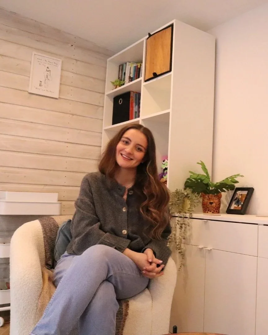 A young woman with long wavy hair, smiling, sitting in an off-white armchair in a cozy room with wooden and white wall paneling, bookshelves, a potted plant, and framed photos.