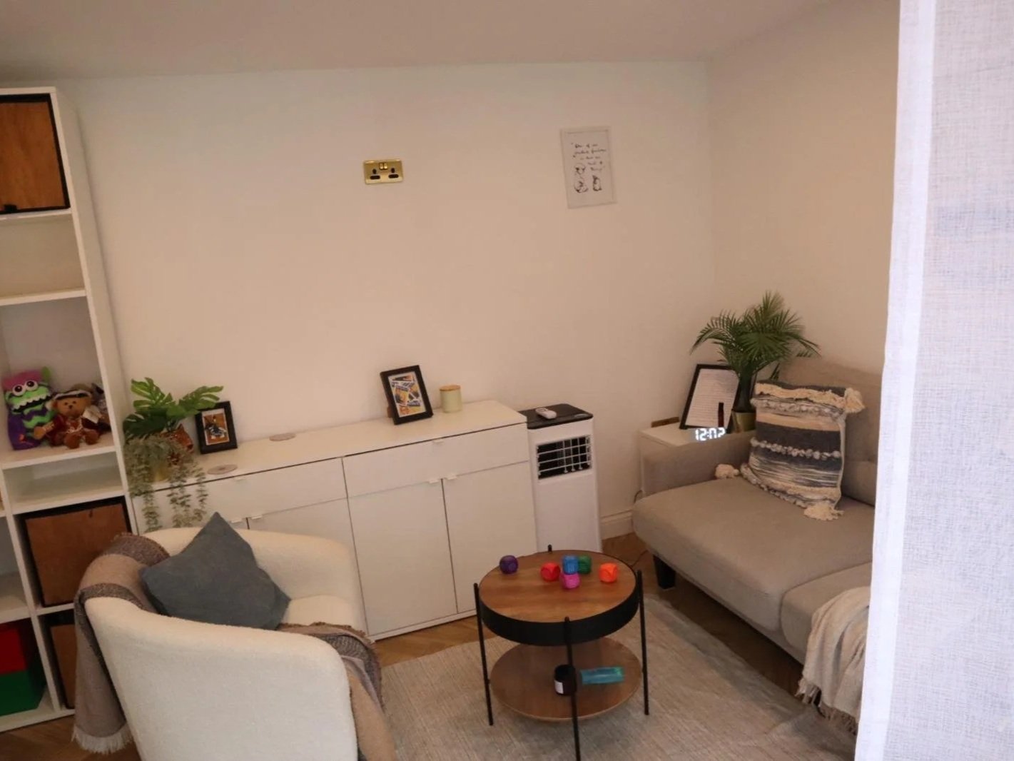 Living room with a beige armchair, a light gray sofa with pillows, a small dark wood coffee table with colorful toys, a white cabinet, a set of shelves with plush toys, potted plant, framed pictures, and a digital clock on a side table showing 12:03.