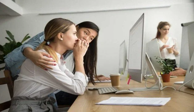 Two women discussing work at an office desk with computers, while a third woman stands in the background.