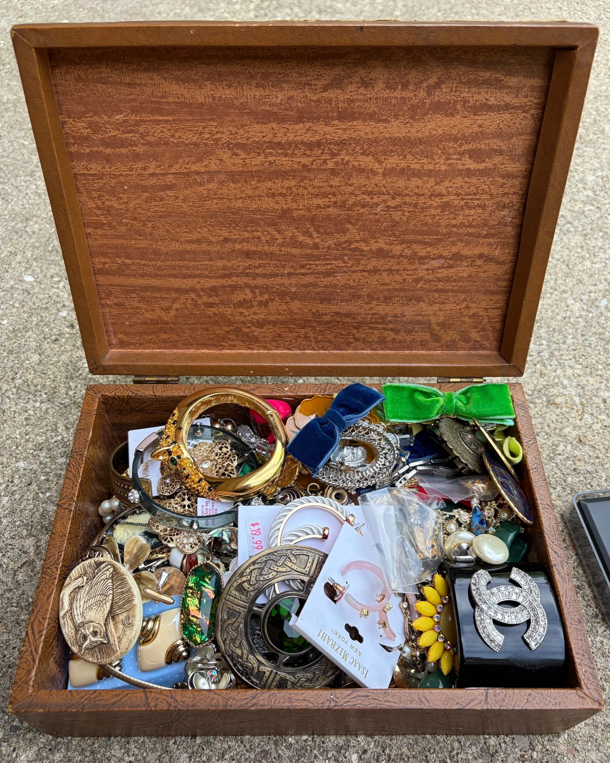 An open wooden jewelry box filled with assorted jewelry including bracelets, earrings, brooches, and watches, some still with tags attached. The jewelry features various styles, colors, and materials, including gold, silver, pearls, and gemstones.