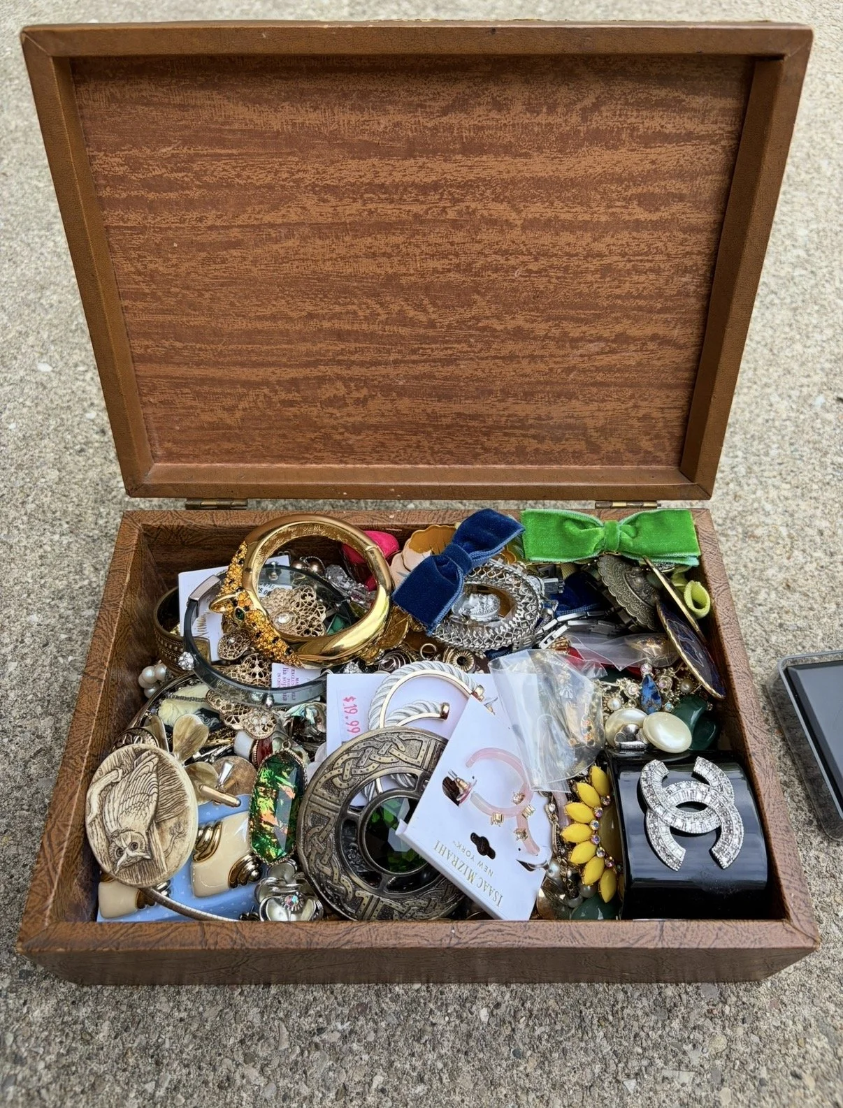 Open wooden box filled with assorted jewelry, including bracelets, earrings, pins, and hair accessories, on a textured surface.