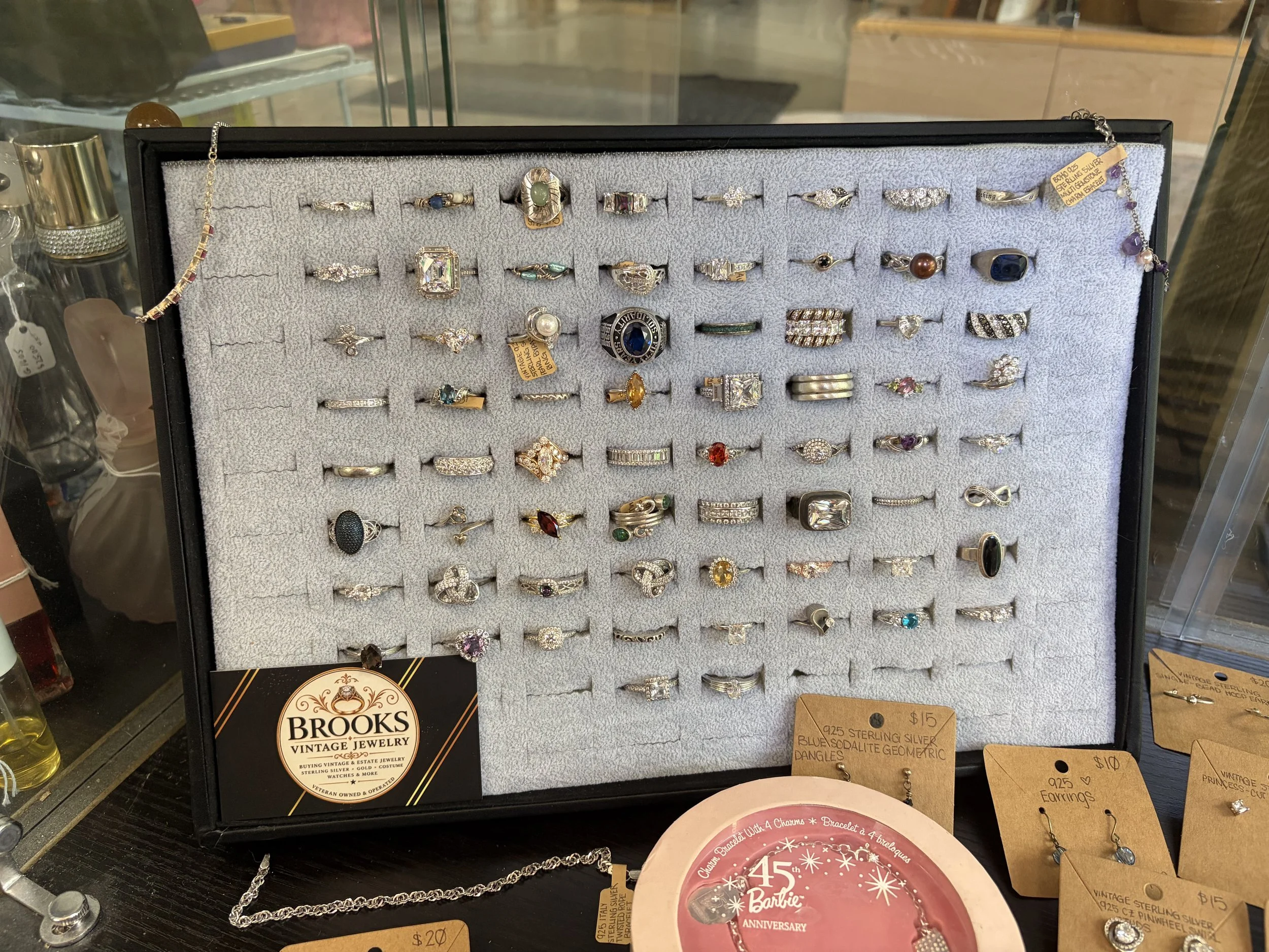 Display case with various rings and jewelry pieces, including rings, earrings, and bracelets, some with large gemstones and intricate designs at Brooks Vintage Jewelry store.