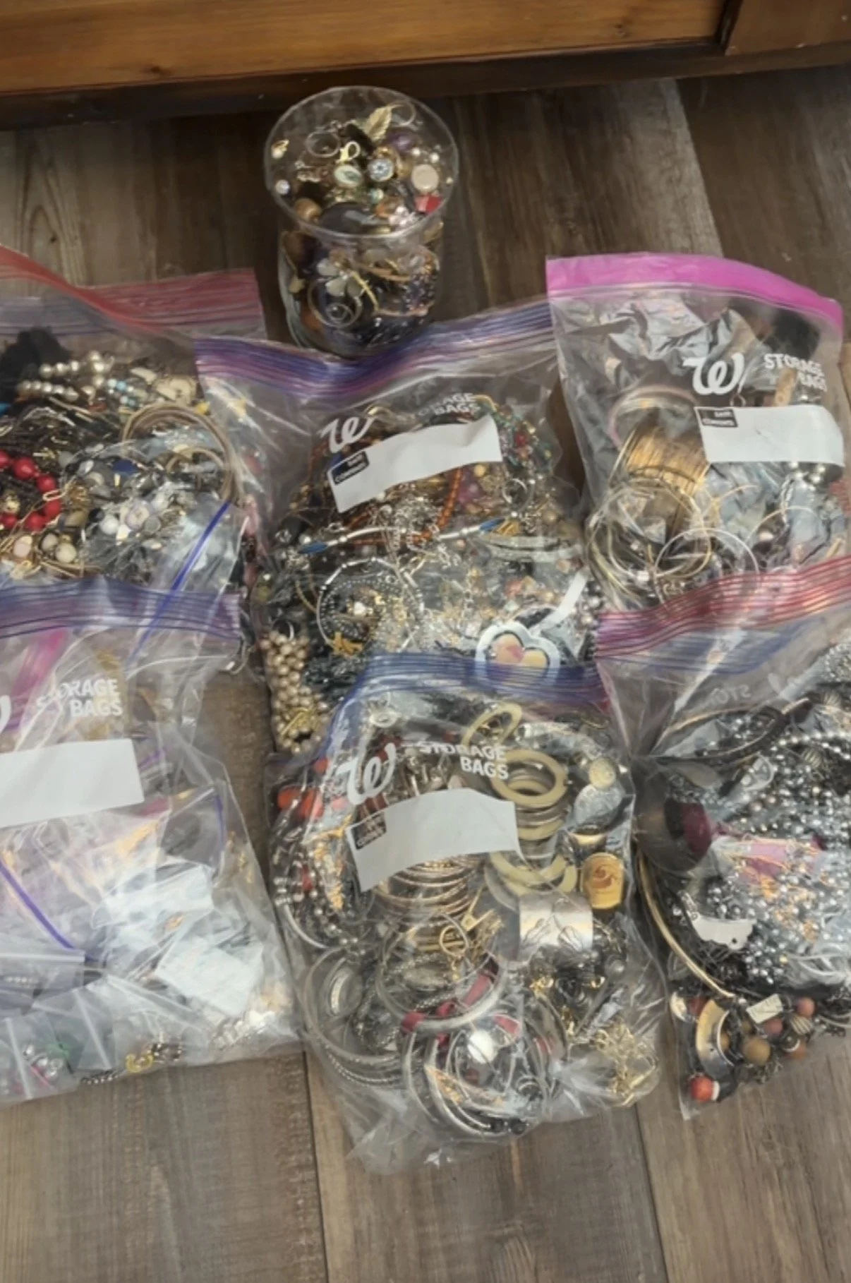 Multiple clear plastic bags filled with assorted jewelry, including earrings, necklaces, bracelets, and rings, on a wooden surface.