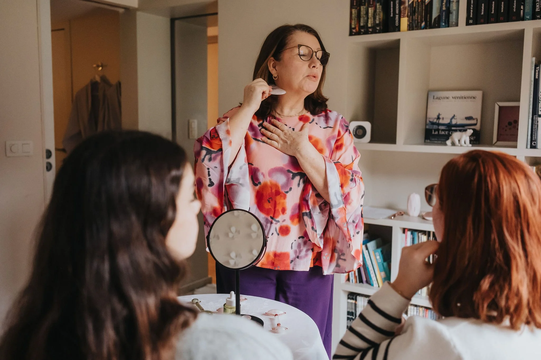 Une femme portant des lunettes et une robe fleuries en train de parler à un groupe de personnes assises autour d'une table dans une bibliothèque ou un salon.