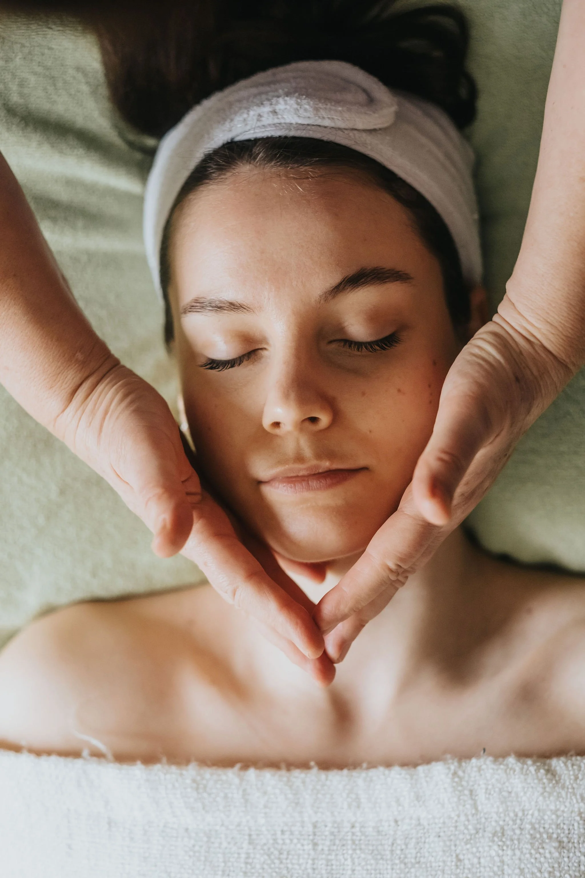 Une jeune fille avec les yeux fermés, un bandeau blanc sur la tête, repose sur un lit et forme un cœur avec ses mains, entourée par une autre paire de mains.