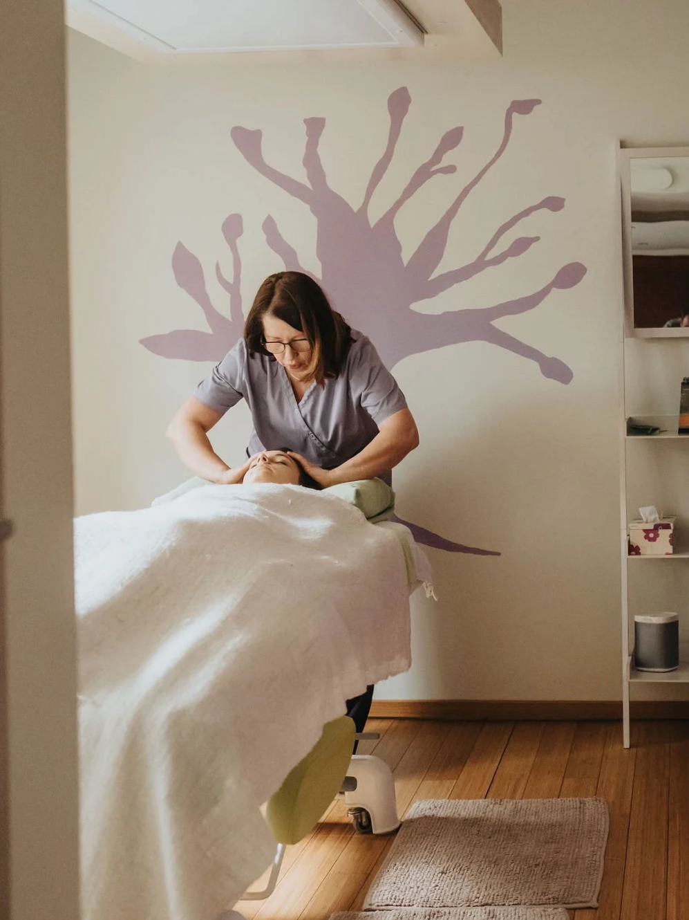 Une femme reçoit un soin de visage ou un massage dans un salon de spa, avec un mur décoré d'une silhouette d'arbre en arrière-plan.