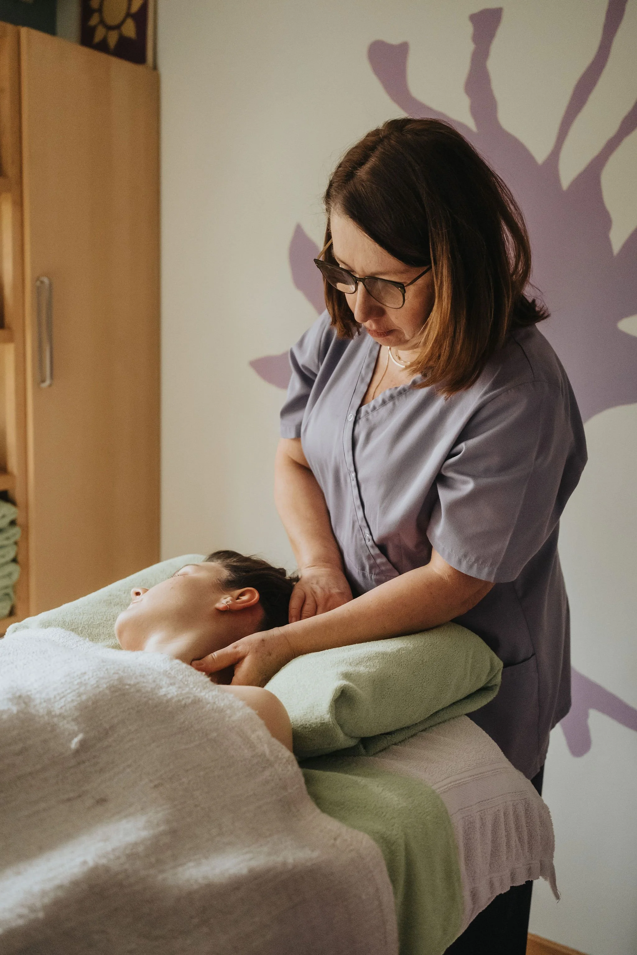 Une femme en blouse grise donne un massage à une autre femme allongée, recouverte d'une serviette blanche, dans une pièce calme avec un mur décoré de motifs végétaux
