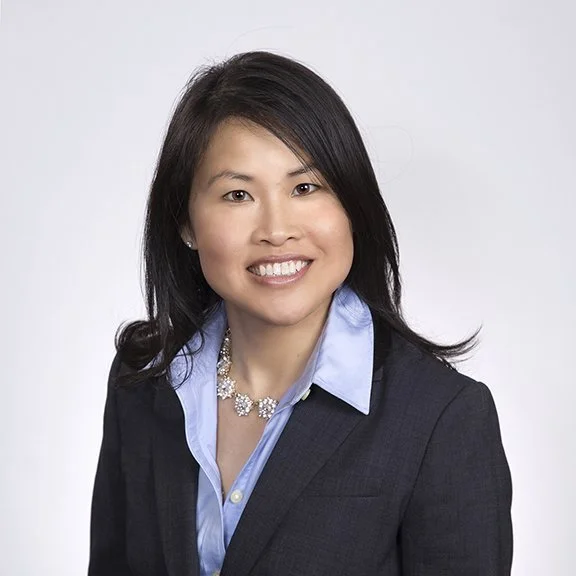 Professional portrait of an Asian woman with black hair, wearing a dark blazer, light blue button-down shirt, and a floral necklace, smiling against a plain white background.