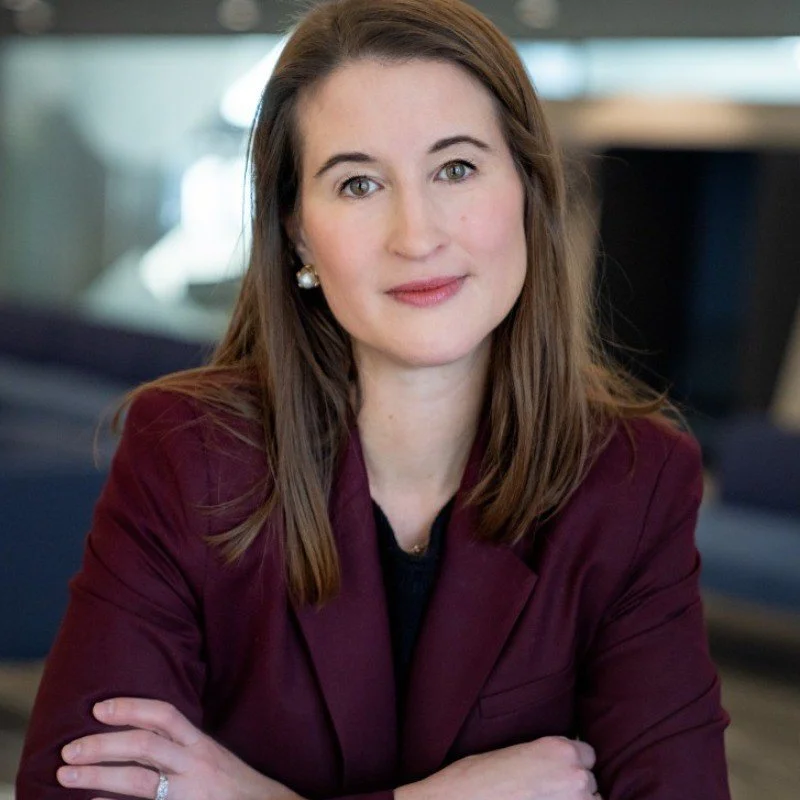 Portrait of a woman with shoulder-length brown hair, wearing a burgundy blazer and pearl earrings, sitting with her arms crossed, in an indoor setting with blurred background.