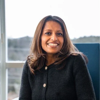 A woman with shoulder-length brown hair and a wide smile, sitting indoors near a window with a blurred outdoor background, wearing a black jacket.