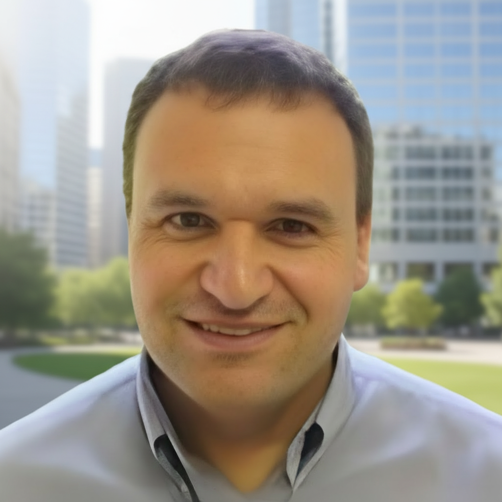 Portrait of a smiling man with short brown hair wearing a light-colored collared shirt, outdoors with modern office buildings and green trees in the background.